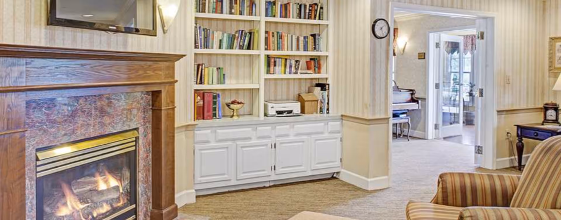 A cozy living room area featuring a lit fireplace with a wooden mantel, built-in white bookshelves filled with books and decorative items, a striped armchair, and a view into an adjacent room with a piano and glass double doors.