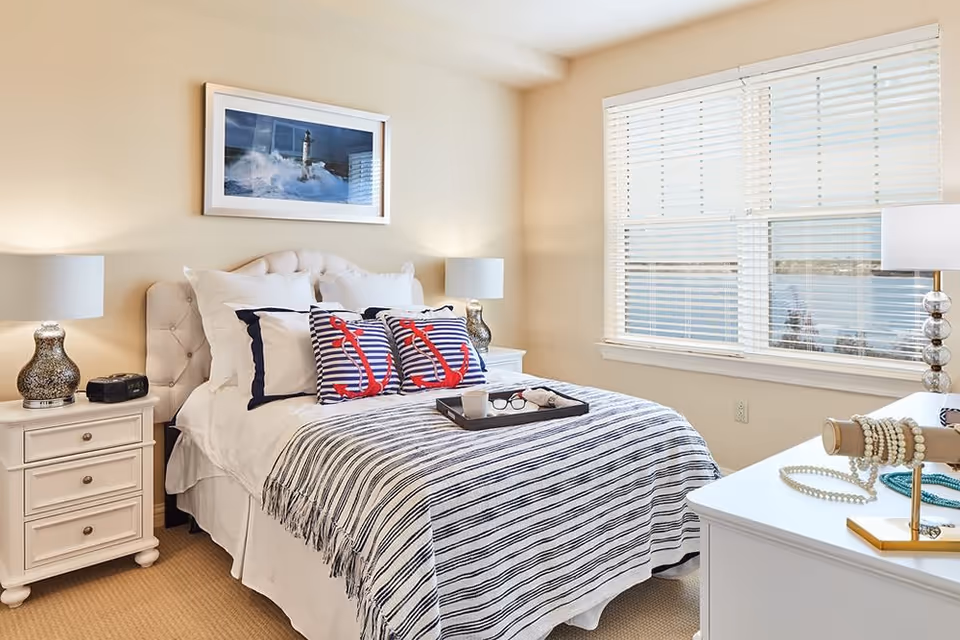 Bright coastal-themed bedroom with a neatly made bed, striped bedding and anchor pillows, nightstands and a window overlooking water.