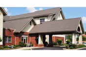 Exterior view of a senior living facility building with a covered entrance, brick and siding walls, and a sloped roof under a blue sky with some clouds.