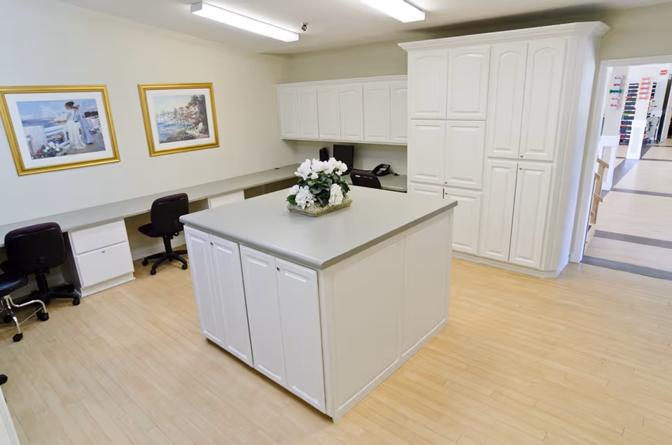 A clean, well-lit office area with white cabinetry and a large central island topped with a flower arrangement. There are several black office chairs along a long desk against the wall, which is decorated with two framed paintings. The floor is light wood, and there is a hallway visible in the background.