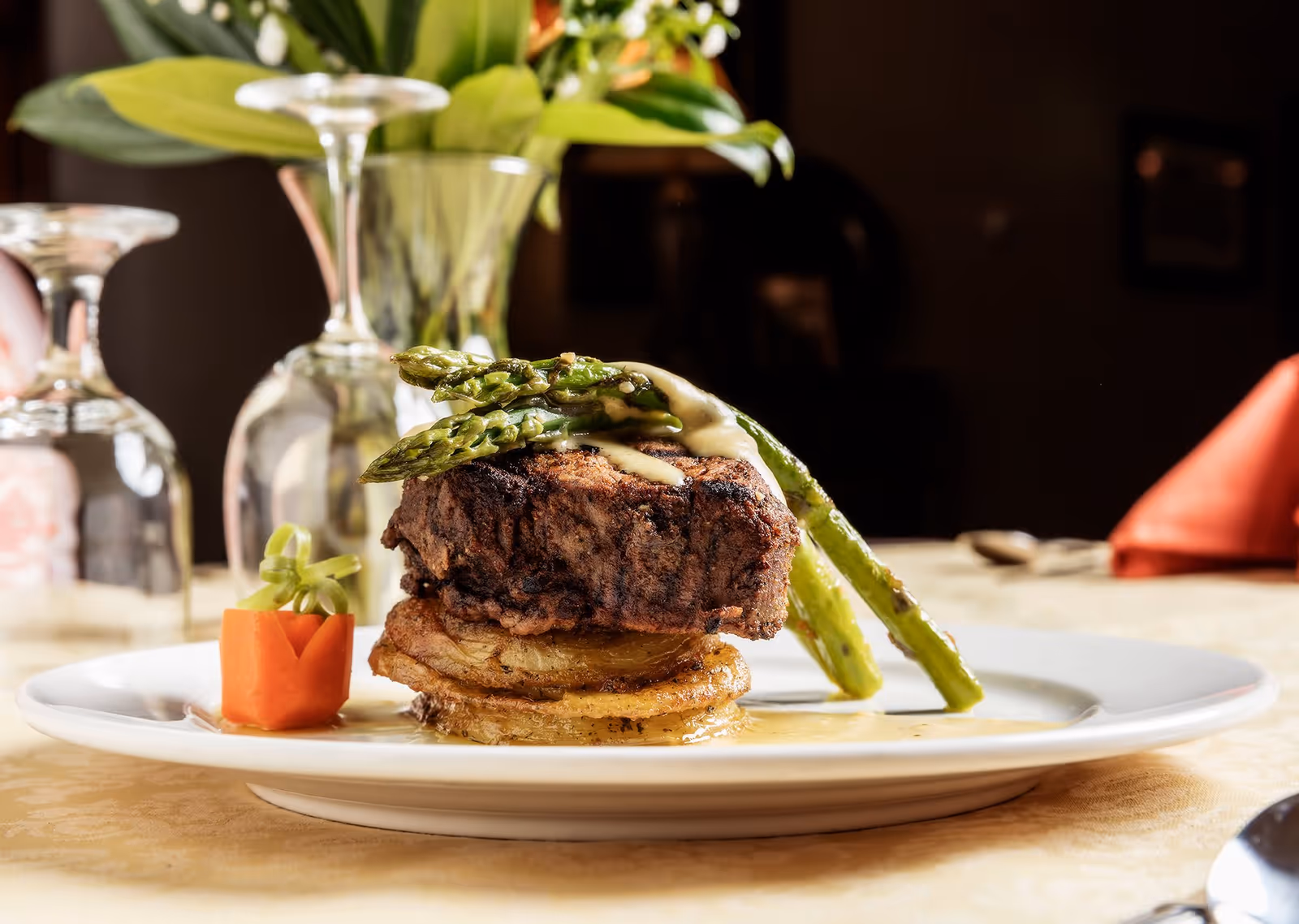 A plated gourmet meal featuring a grilled steak topped with asparagus and sauce, served on a bed of sliced potatoes with a small carved carrot garnish, set on a table with a beige tablecloth and glassware in the background.