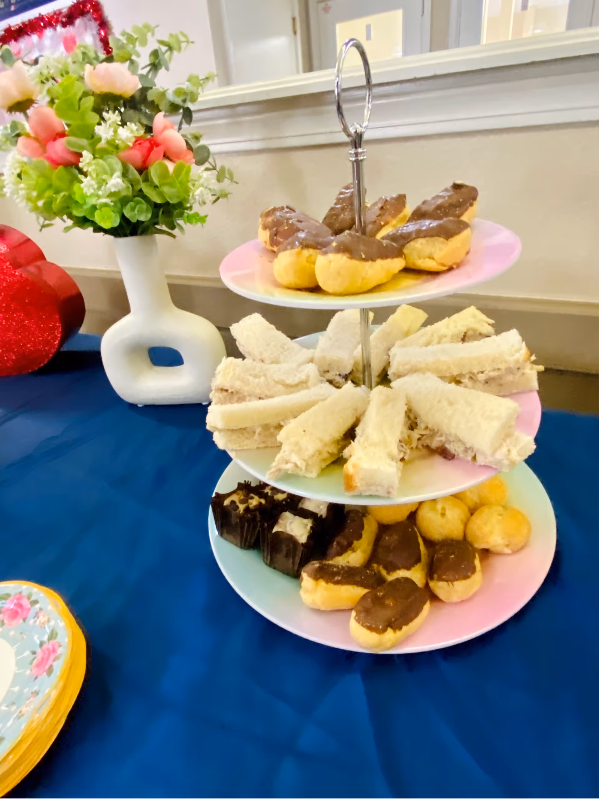 Three-tiered serving tray with sandwiches and chocolate-topped pastries on a blue tablecloth next to a vase of flowers.