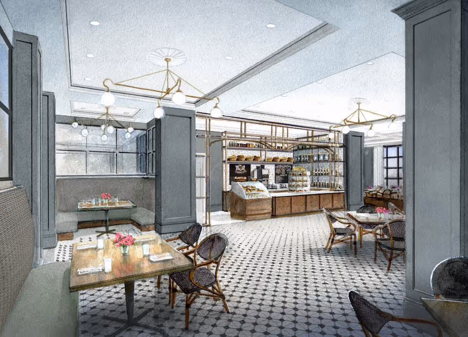 Interior view of a dining area with tables and chairs arranged neatly. The room features patterned tile flooring, gray walls with paneling, and modern light fixtures hanging from the ceiling. In the background, there is a counter area with shelves displaying baked goods and a menu board.