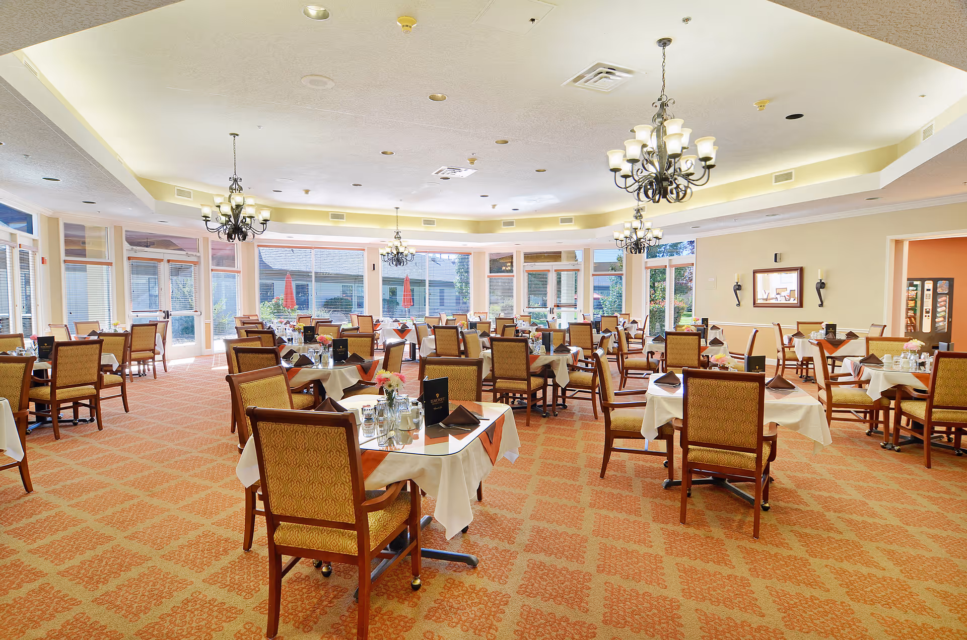 Spacious dining room with multiple set tables and chairs, chandeliers, and large windows looking out to an outdoor patio.