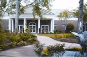 Front entrance of a brick building with white columns and a covered portico, a curved walkway, landscaped planting beds and a small fountain sculpture.