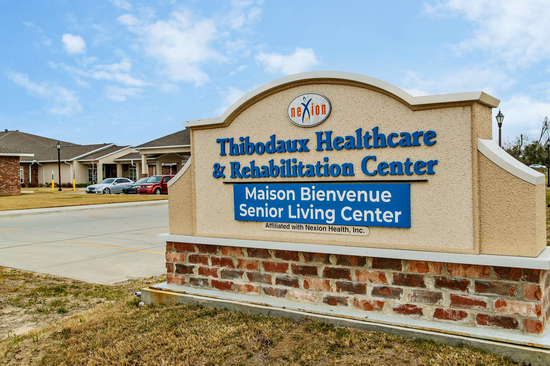 Outdoor view of the entrance sign for Thibodaux Healthcare & Rehabilitation Center and Maison Bienvenue Senior Living Center, with the facility building and parked cars visible in the background under a partly cloudy sky.