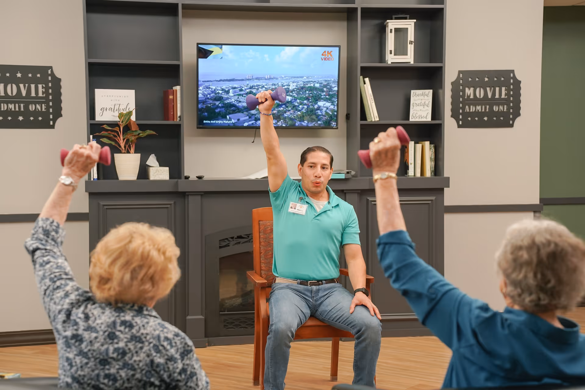 A man in a teal shirt leads a seated exercise class for two elderly women, all lifting pink dumbbells in a room with a TV mounted on the wall and decorative shelves.