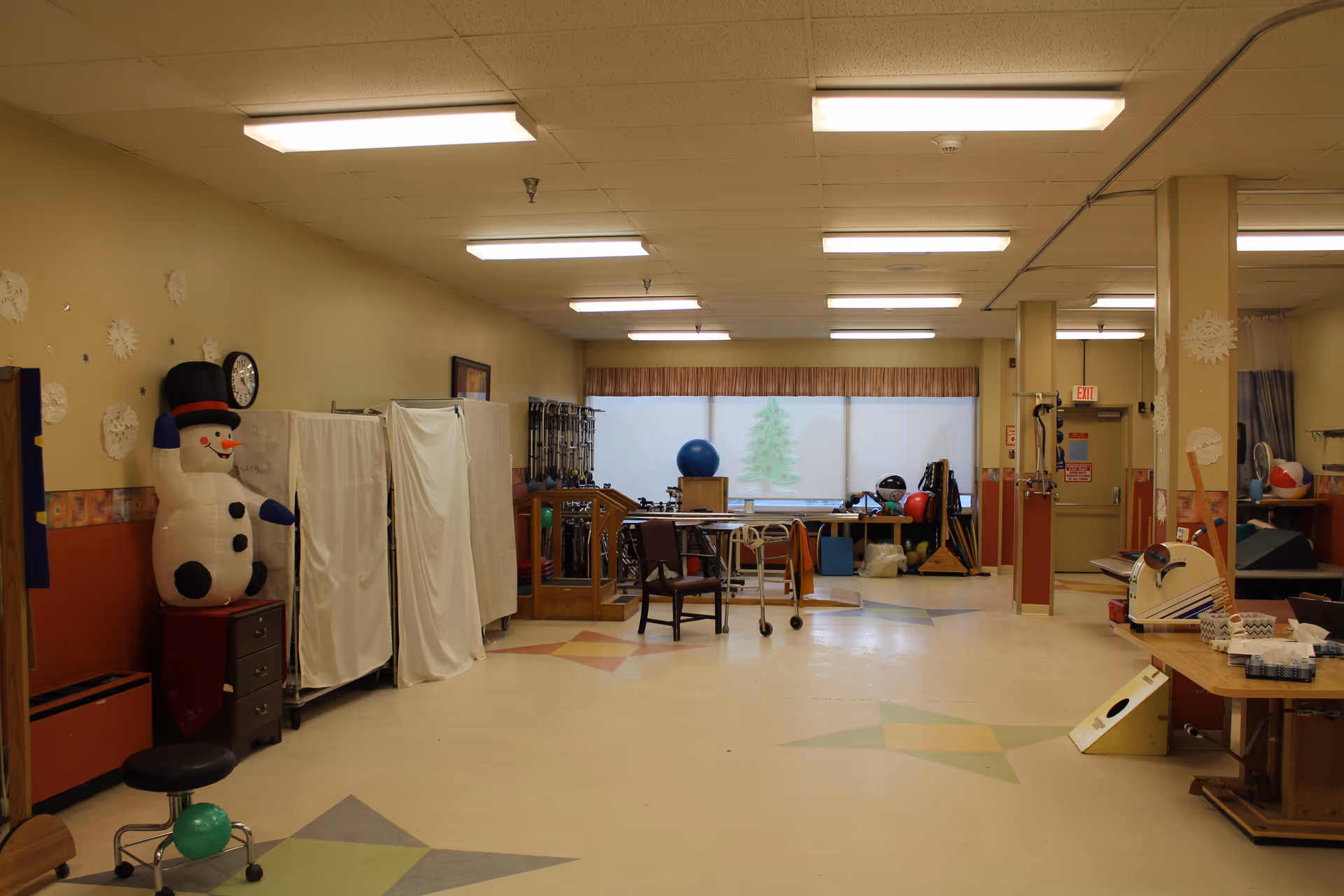 A spacious room with beige and red walls, decorated with paper snowflakes and a large inflatable snowman. The room contains various exercise and therapy equipment, including chairs, walkers, exercise balls, and therapy tables. The floor has colorful star patterns, and fluorescent lights illuminate the ceiling. A window at the far end has a frosted design with a tree image.