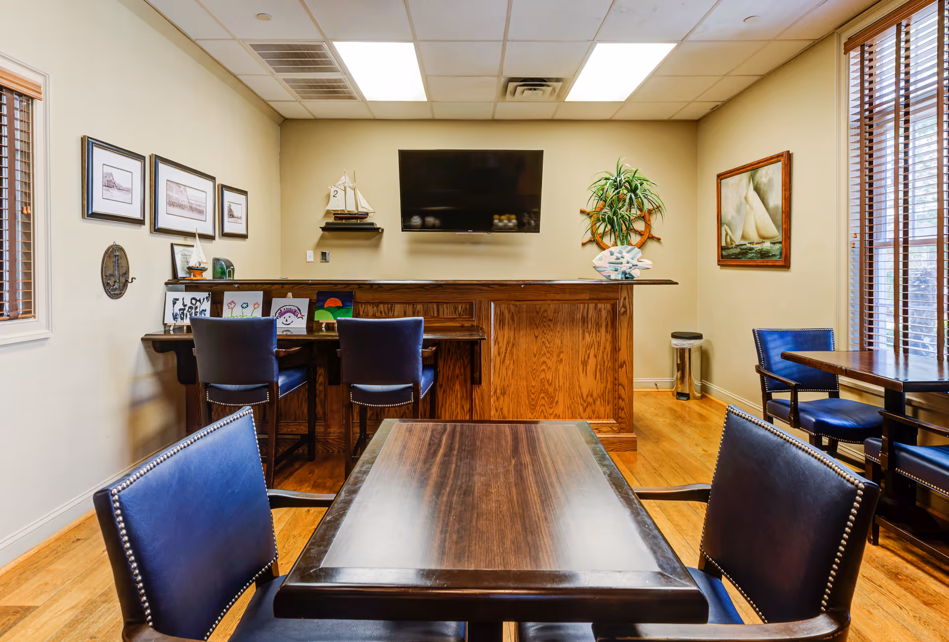 Interior view of a room with wooden floors and beige walls featuring a wooden table with four blue leather chairs in the foreground. In the background, there is a wooden counter with two blue chairs, a flat-screen TV mounted on the wall, framed pictures, a small model sailboat, and a plant with a ship's wheel decoration. Large windows with wooden blinds allow natural light into the room.