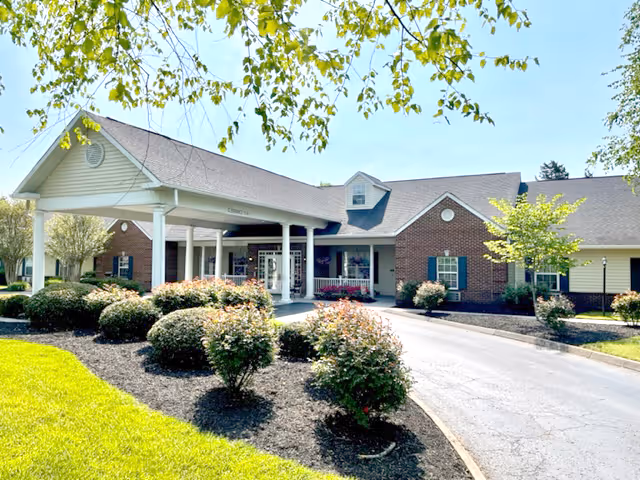 Front entrance of a brick senior living building with a covered portico, circular driveway, and landscaped shrubs.