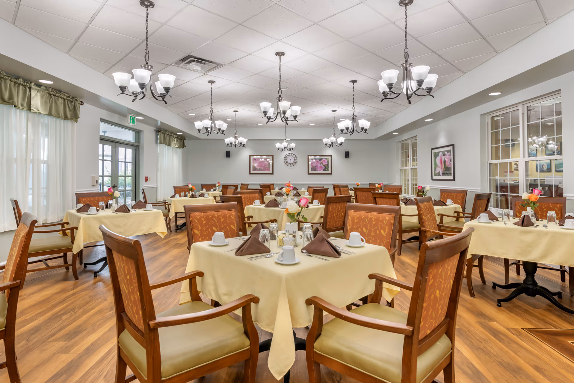 Bright, spacious dining room with multiple tables set with yellow tablecloths, wooden chairs, chandeliers, and floral centerpieces.