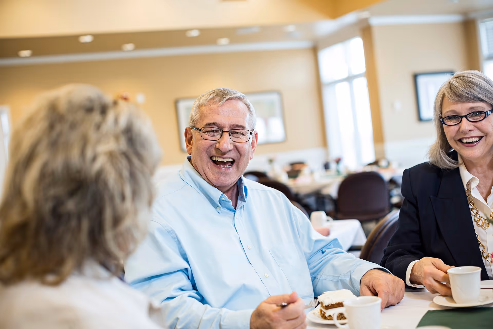 Three seniors sitting at a table in a dining area, smiling and enjoying coffee and cake together. The room has beige walls, large windows, and several tables and chairs in the background.