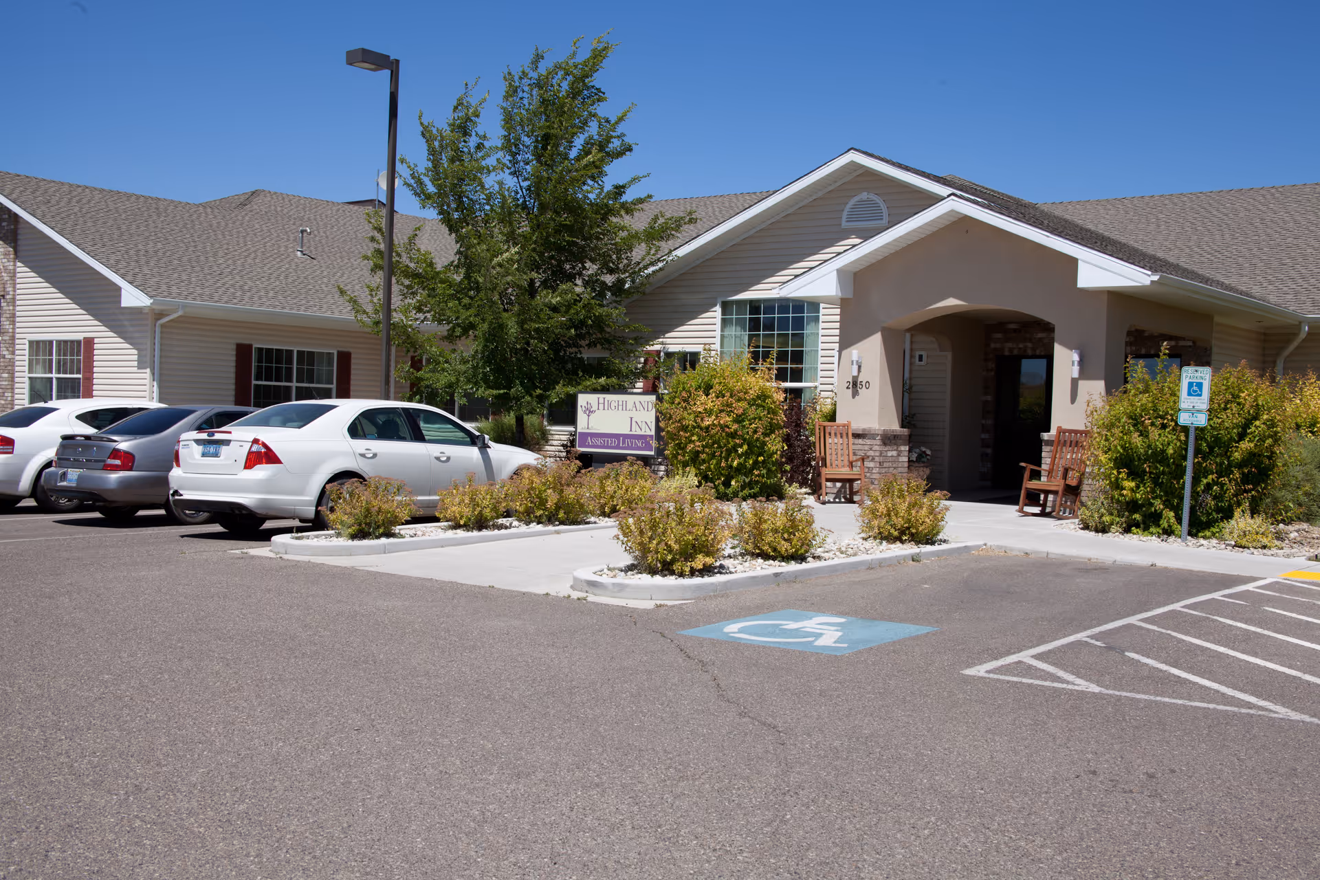 Exterior view of Highland Village of Elko, showing the entrance to the building with a covered porch, two wooden chairs, bushes, and a parking lot with several parked cars including a handicapped parking space.