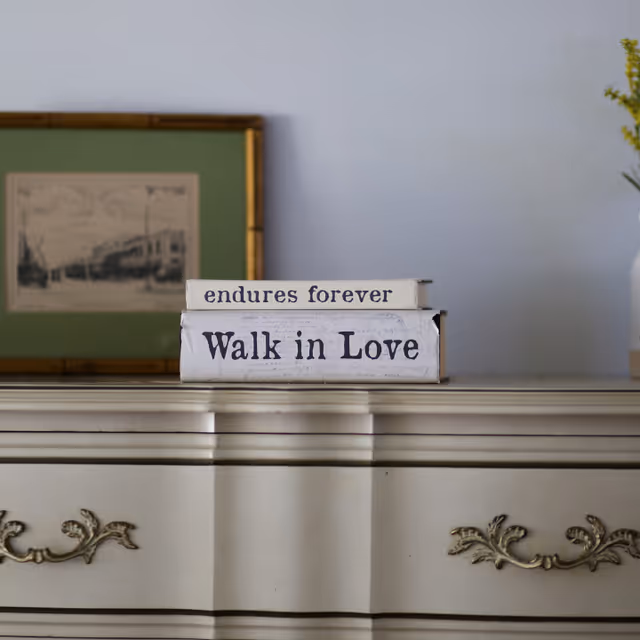 Two decorative books stacked on a white vintage dresser. The top book's spine reads 'endures forever' and the bottom book's spine reads 'Walk in Love'. In the background, there is a framed black and white picture and a vase with yellow flowers on the right side.