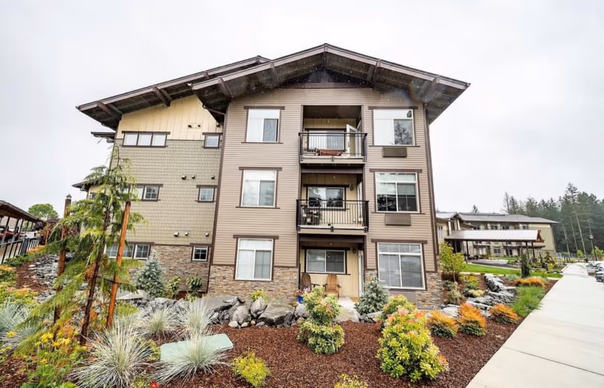 Exterior view of a three-story residential building with balconies and windows, surrounded by landscaped gardens with various plants and shrubs, and a paved walkway on the right side leading to other buildings in the background under an overcast sky.
