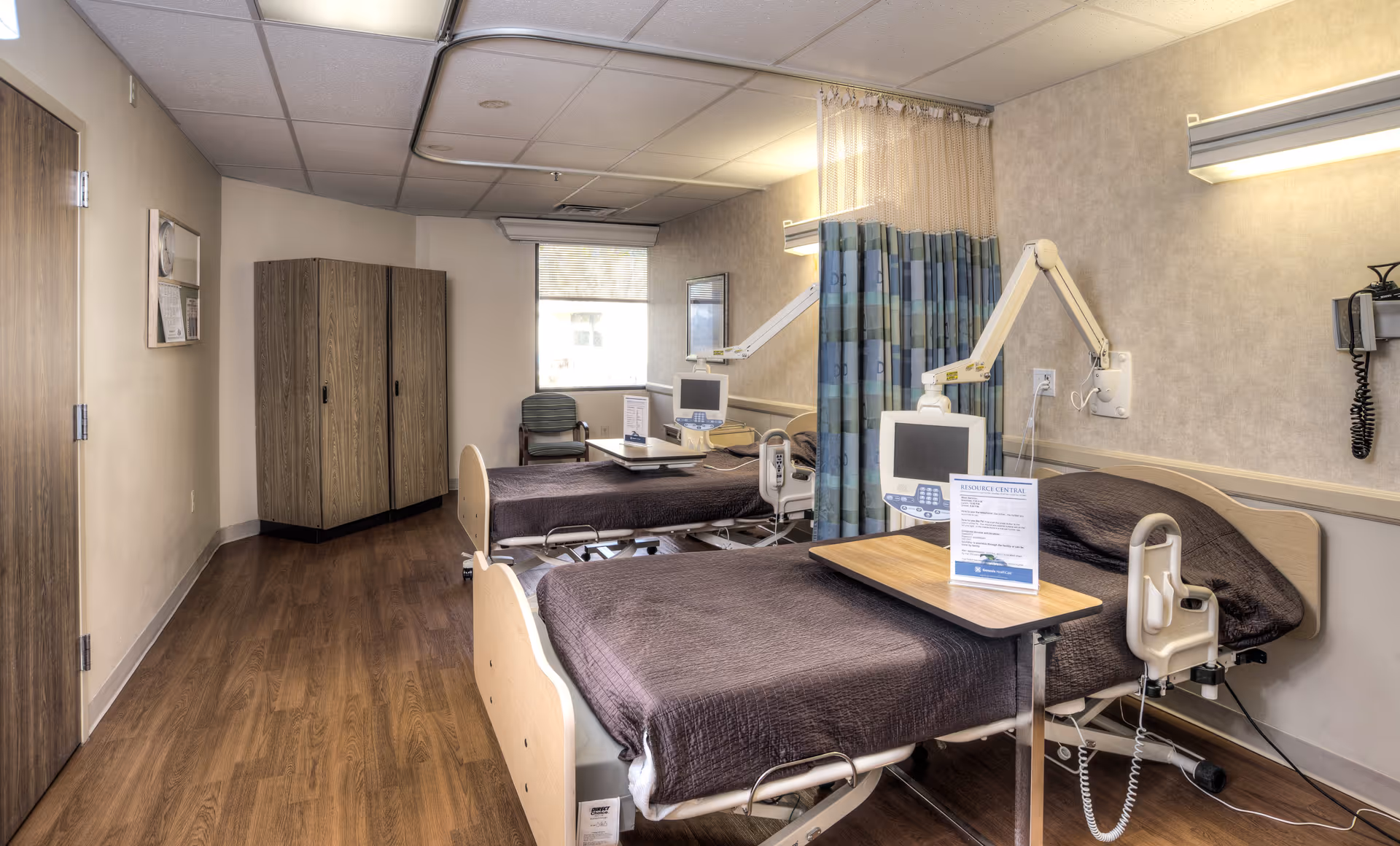 Two hospital-style beds in a rehabilitation room with medical equipment, a privacy curtain, a wardrobe, and wood-look flooring.