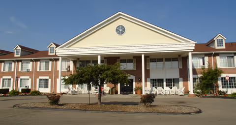 Front exterior view of a two-story senior living facility building with a large covered entrance supported by white columns. The building has a brick and beige siding facade with multiple windows and a circular window at the top of the entrance. There are white chairs arranged on the porch and a small tree and shrubs in front of the building under a clear blue sky.