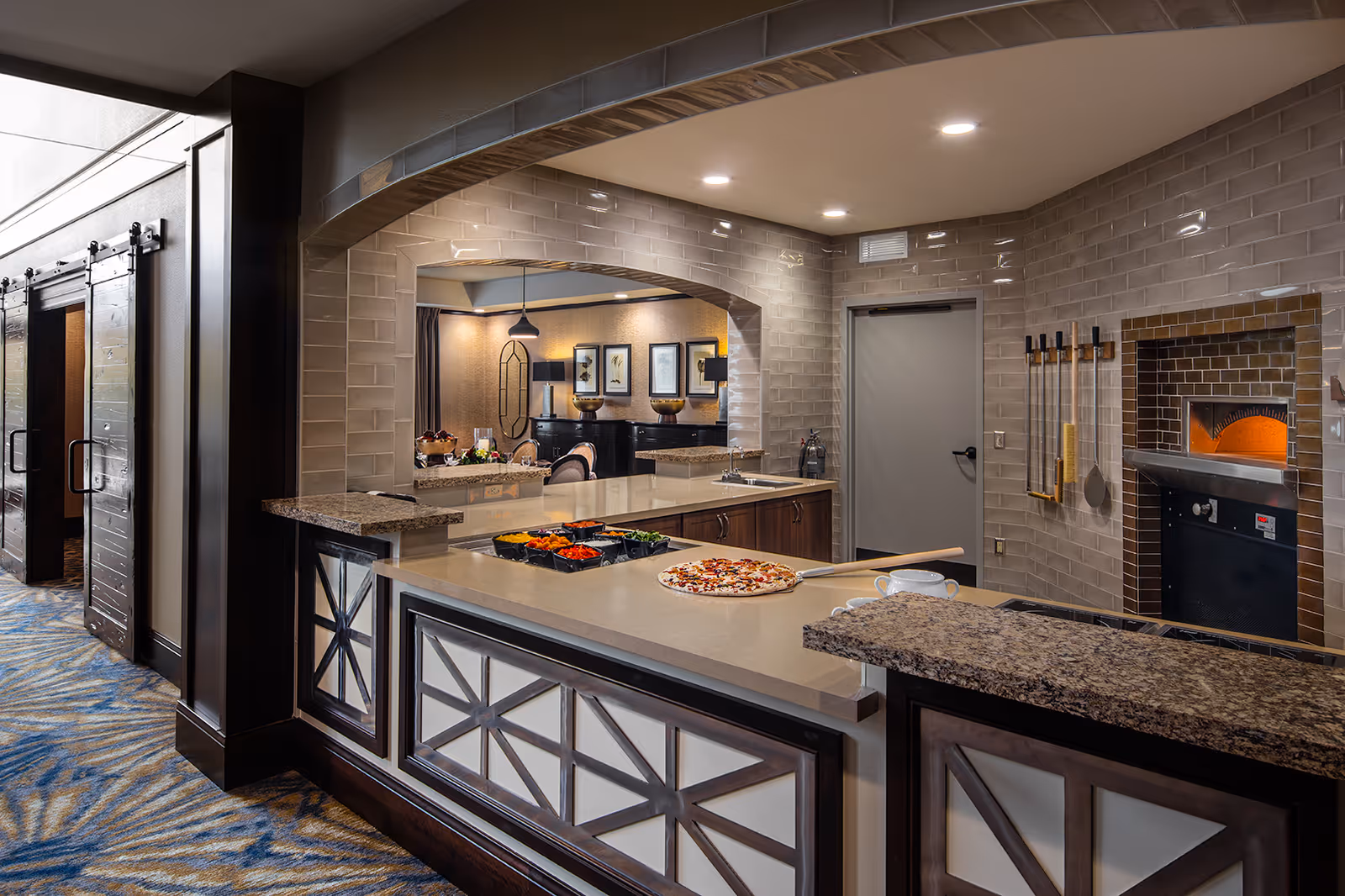Modern kitchen area with a pizza oven built into a tiled wall, a countertop with pizza ingredients and a pizza peel holding a freshly made pizza. The kitchen has a granite countertop and a pass-through window looking into a dining or living area with framed pictures on the wall.
