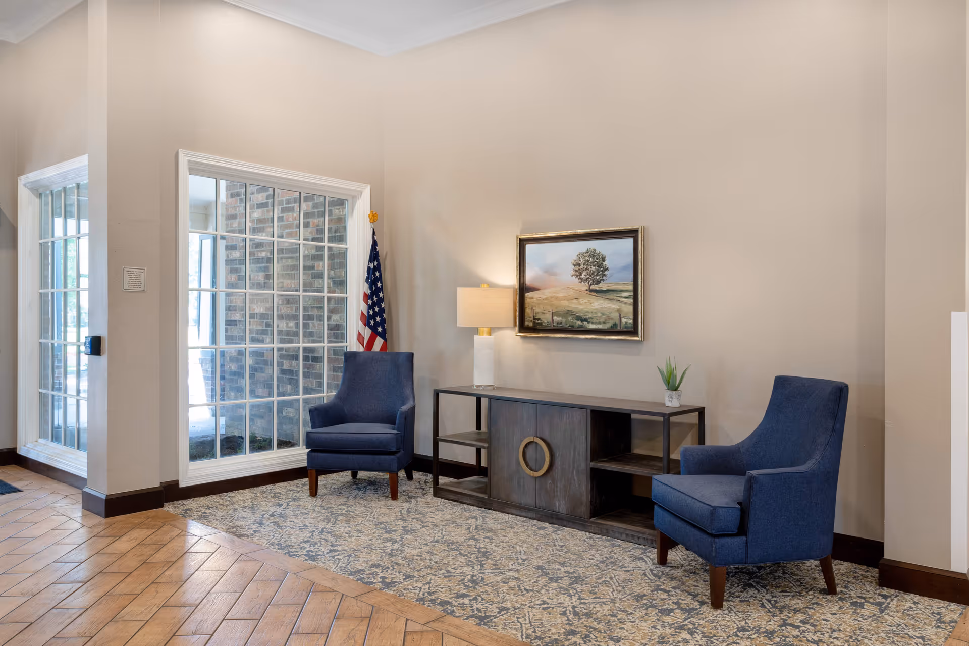 A seating area in a senior living facility featuring two blue upholstered armchairs on a patterned rug, a dark wooden console table with a lamp, a small plant, and a framed landscape painting on the wall. There is a large window with white framing showing a brick exterior wall and an American flag placed between the window and one of the chairs.