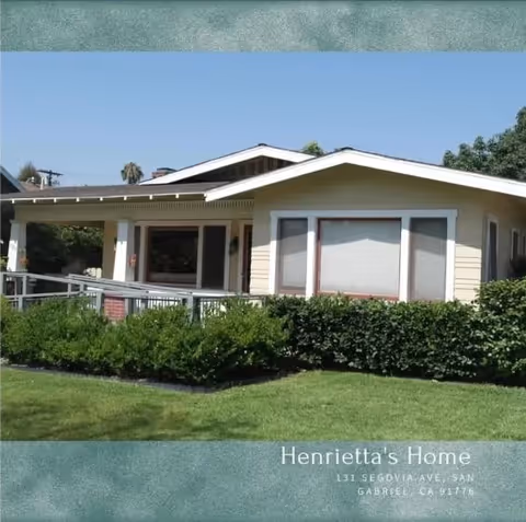 Exterior view of a single-story residential building with a covered porch, surrounded by green bushes and a well-maintained lawn under a clear blue sky.
