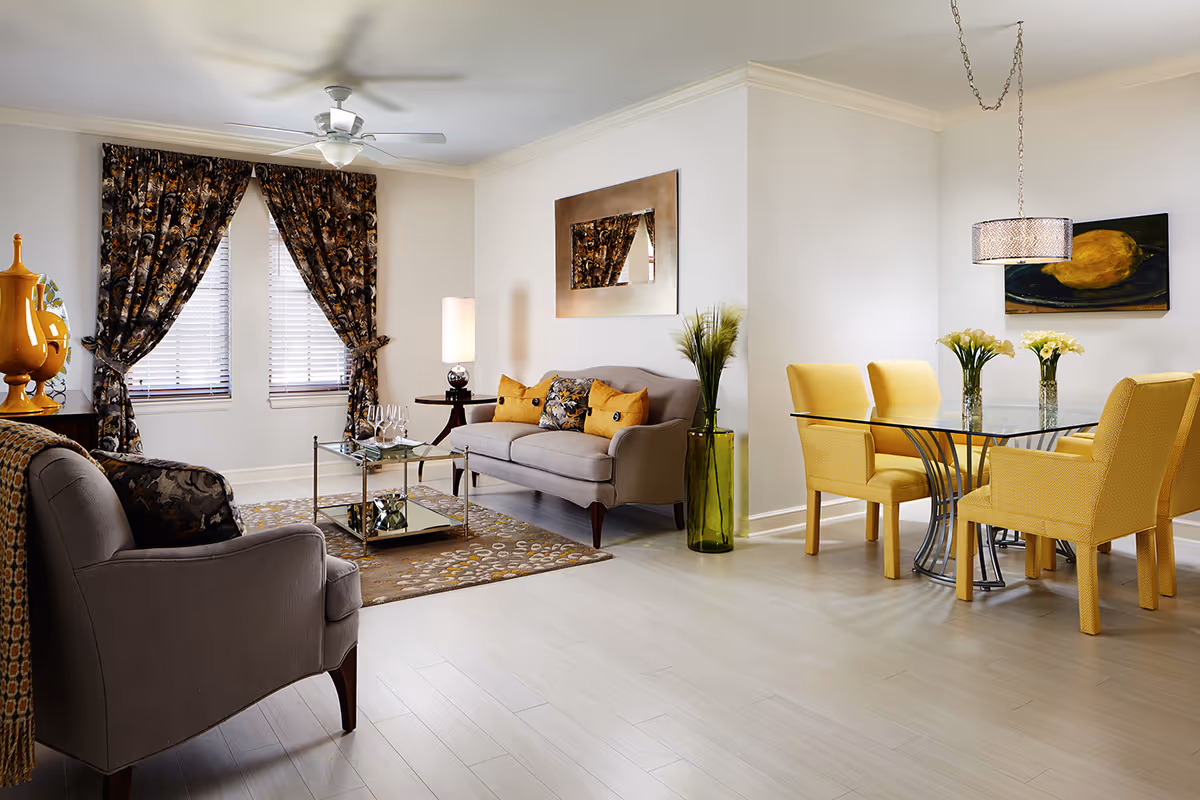 Bright open living and dining area with gray sofas, a glass coffee table, yellow dining chairs around a glass table, patterned curtains, and decorative accents.
