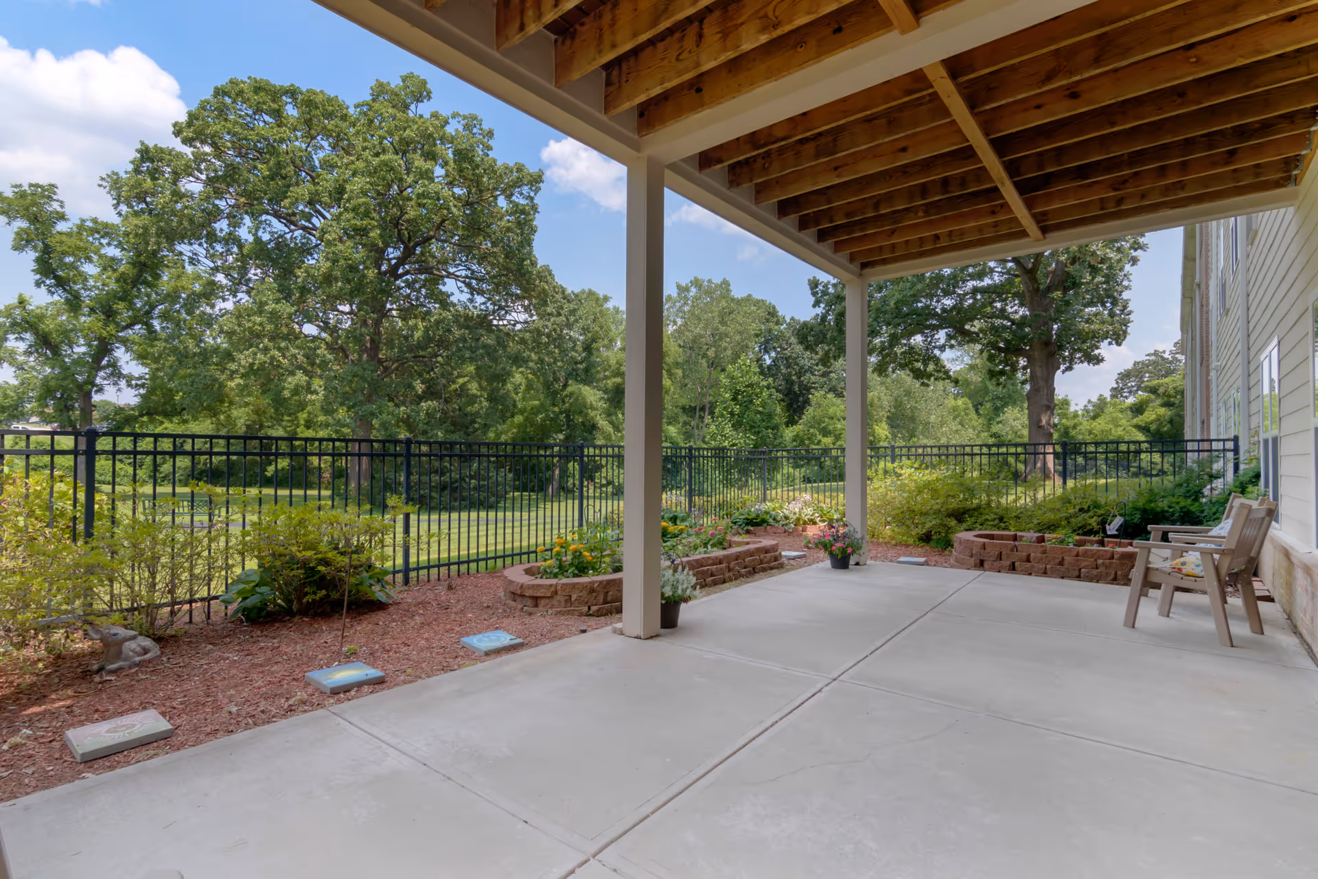 Covered outdoor patio area with concrete flooring, wooden ceiling beams, and two chairs with cushions. The patio overlooks a fenced garden area with various plants, flowers, and large green trees under a partly cloudy sky.
