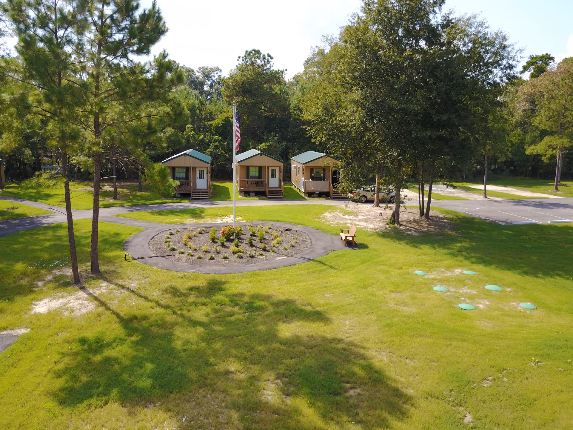 Aerial view of a grassy courtyard with a circular flower bed and flagpole in front of three small cabins surrounded by trees.