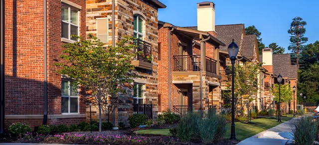 Exterior view of a senior living facility with brick and stone buildings, balconies, trees, shrubs, and a sidewalk lined with street lamps under a clear blue sky.