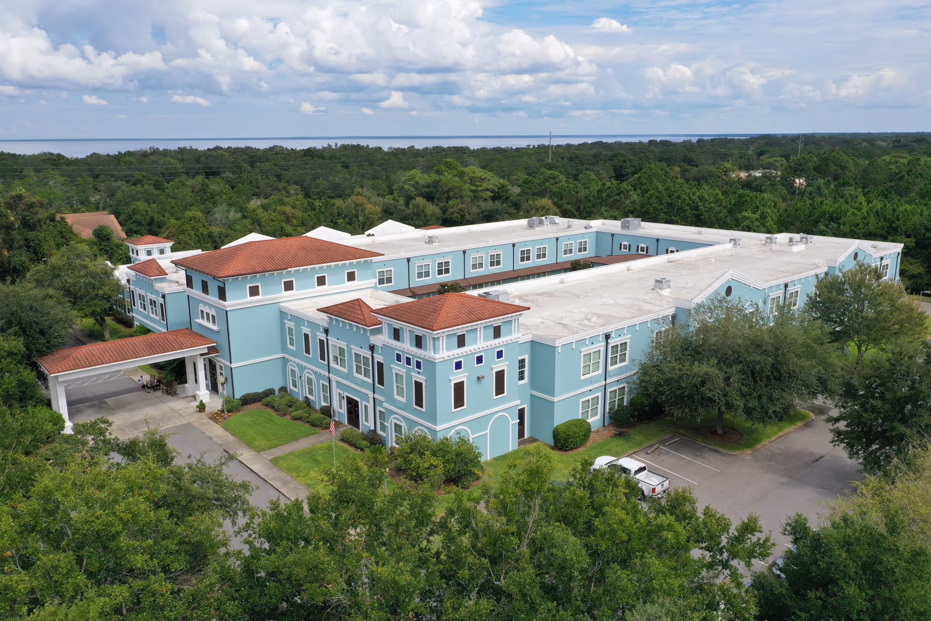 Aerial view of a large, light blue senior living facility building with red-tiled roofs surrounded by green trees and a parking area. The building has multiple windows and a covered entrance driveway. The background shows a forested area and a body of water under a partly cloudy sky.