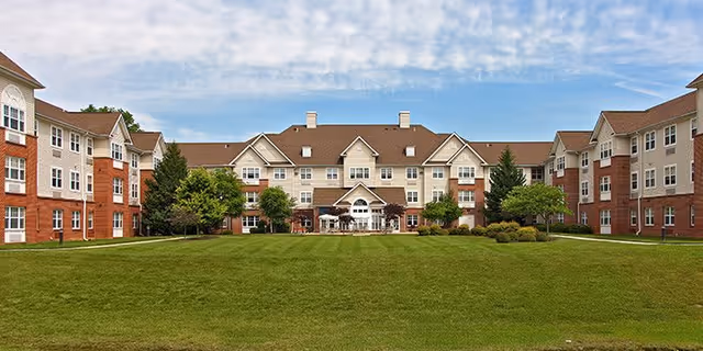 Wide exterior view of Hampshire Village senior living facility showing a large, well-maintained green lawn in the foreground and a multi-story building with brick and beige siding in the background under a partly cloudy sky.