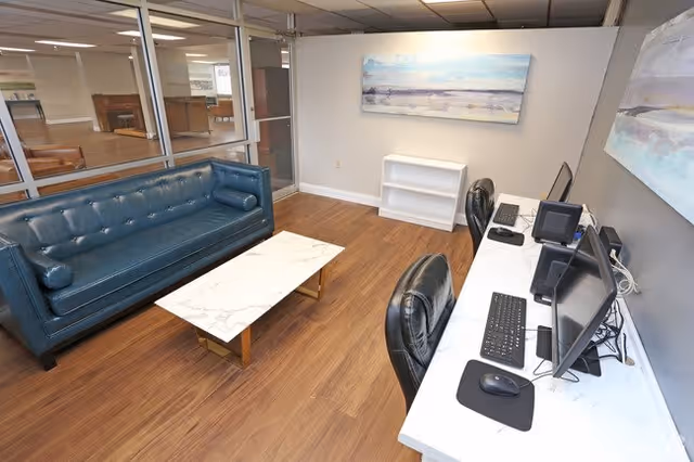 Interior view of a modern office or common area with a blue leather tufted sofa, a white marble coffee table with gold legs, and a white desk with two black office chairs and computer monitors. The room has wood flooring, a large window with a view into another room, and a painting of a beach scene on the wall.