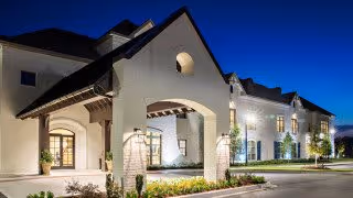Exterior view of The Blake at Chenal Valley facility at dusk, showing a well-lit entrance with an arched portico, white walls, and a dark roof under a clear blue sky.