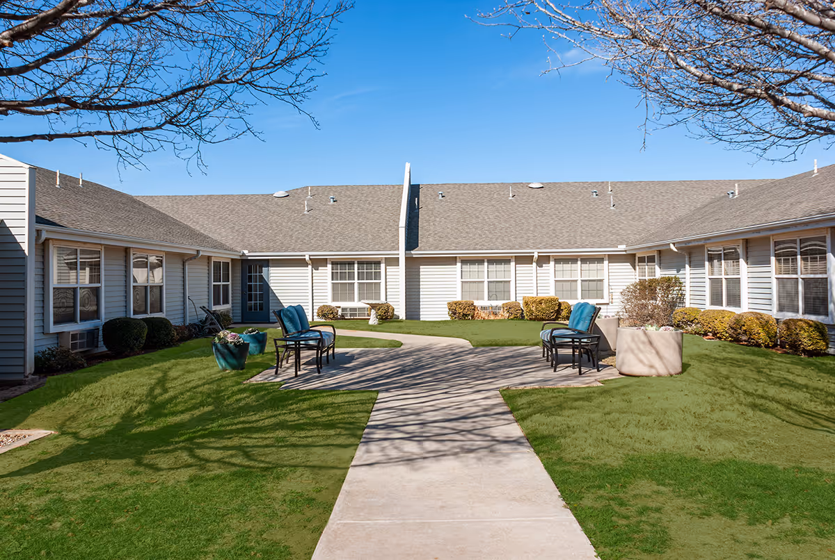Outdoor courtyard area at Cimarron Place with a concrete walkway leading to a seating area featuring chairs with blue cushions and large planters. The courtyard is surrounded by a single-story building with multiple windows and trimmed bushes. Leafless tree branches frame the top of the image under a clear blue sky.