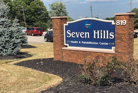 Outdoor view of the entrance sign for Seven Hills Health & Rehabilitation Center, mounted on a brick structure with the number 819. The sign is surrounded by landscaping including bushes and a pine tree, with a parking lot and cars visible in the background under a partly cloudy sky.