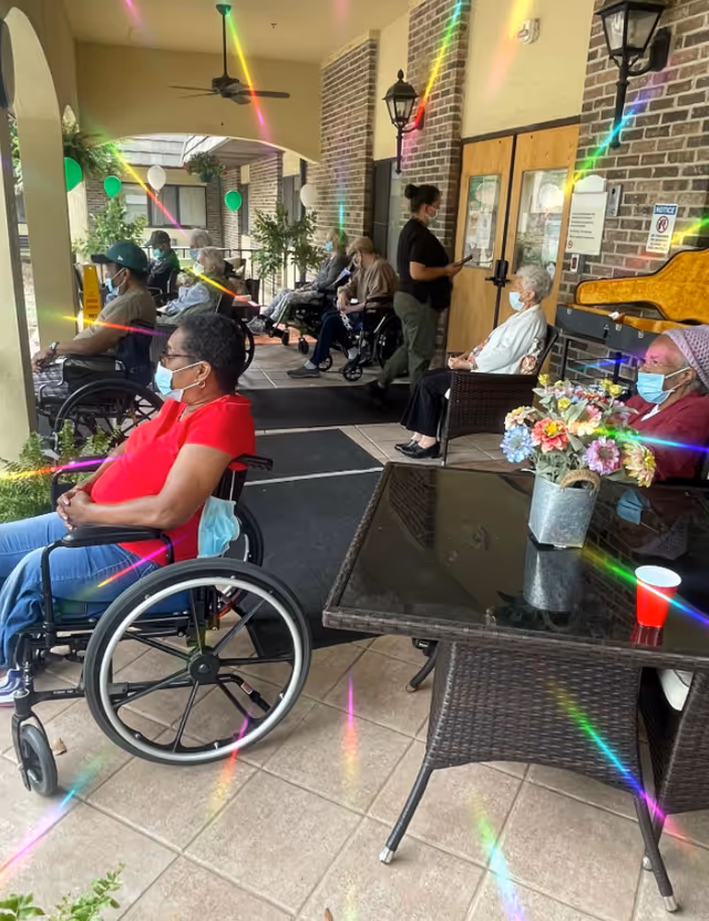 Several elderly individuals, most in wheelchairs and wearing face masks, are seated on a covered outdoor patio area with tiled flooring and brick walls. A caregiver is standing near a door, interacting with one of the residents. The patio is decorated with green and white balloons and potted plants. There is a glass-top table with a flower arrangement and a red cup in the foreground.