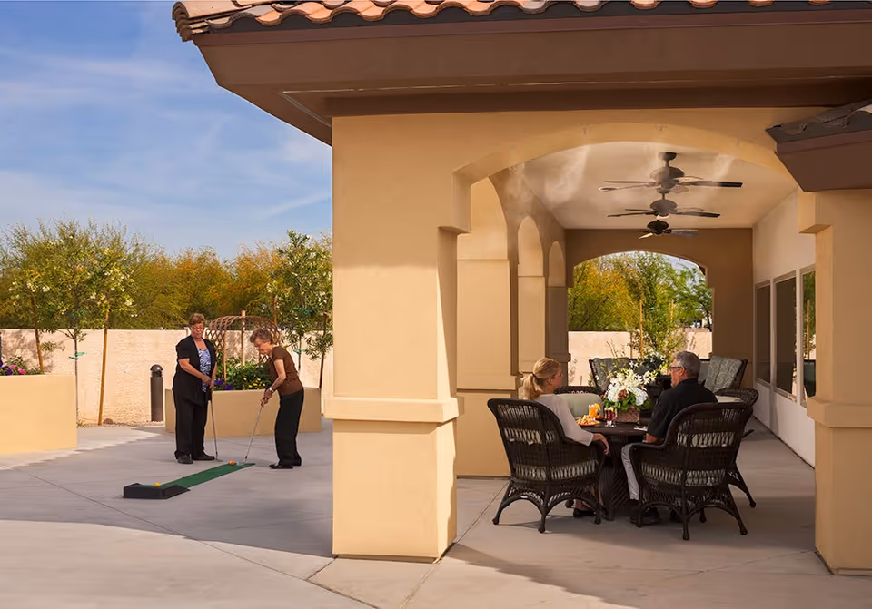Two women playing mini golf on a putting green outdoors near a beige stucco building with arches. Nearby, a man and a woman are sitting and talking at a round table with wicker chairs under a covered patio with ceiling fans. Trees and a clear blue sky are visible in the background.