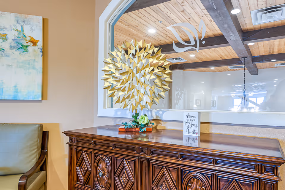 Ornate wooden sideboard topped with a gold sunburst sculpture, a small decorative sign, and a mirror reflecting a beamed ceiling in a communal interior.