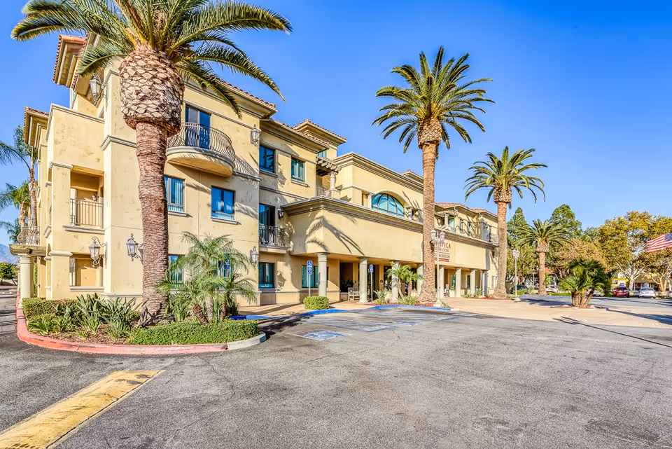 Exterior view of a three-story beige building with balconies and large palm trees in front, surrounded by a parking lot with handicap parking spaces and clear blue sky.
