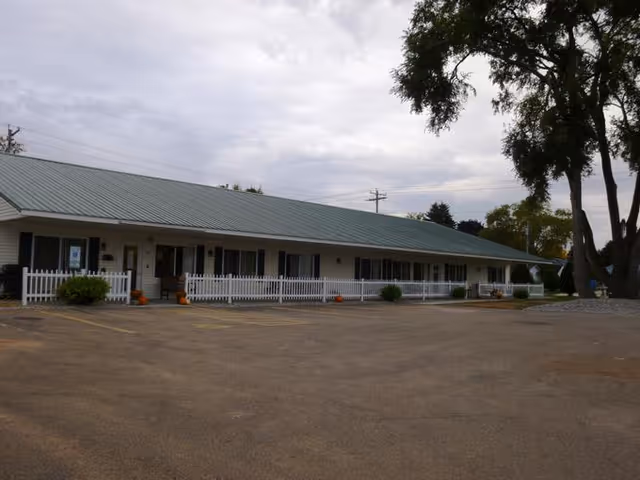 Single-story building with a green metal roof and white exterior, featuring a white picket fence along the front. There are several windows and doors visible, with some small bushes and pumpkins near the entrance. The building is surrounded by a large paved parking area and some trees are visible on the right side under a cloudy sky.