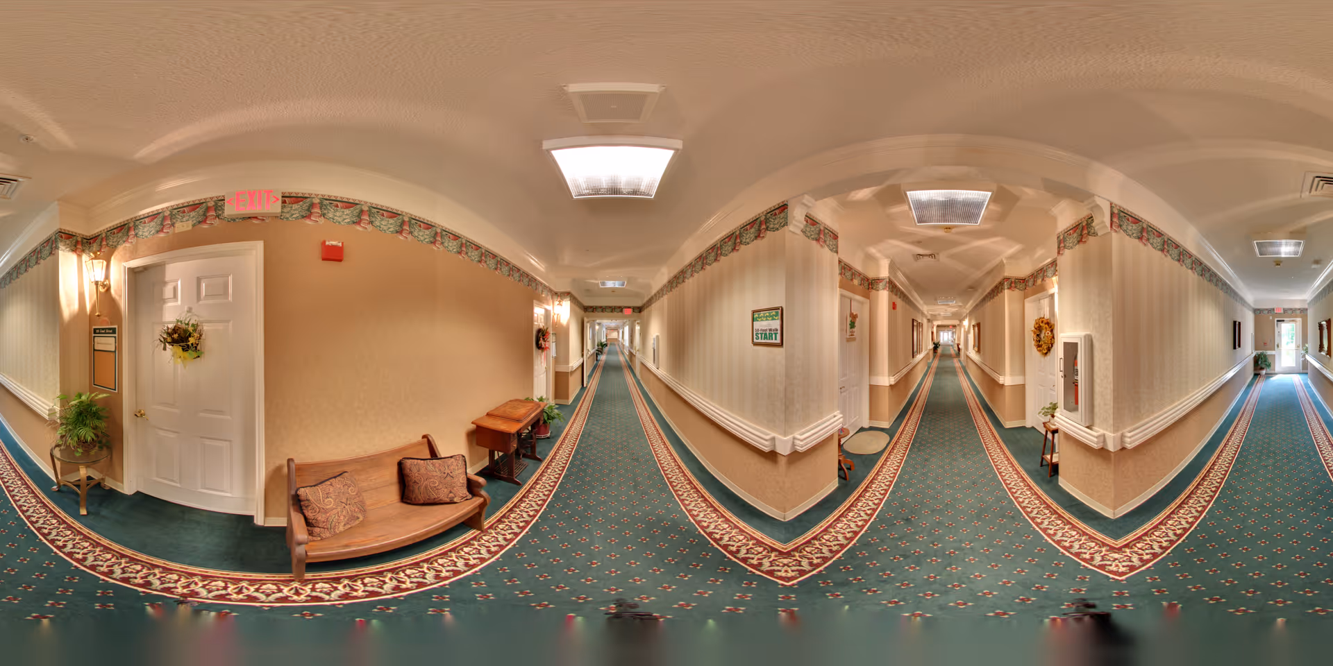 A long, carpeted hallway in a senior living facility with beige walls and decorative wallpaper border near the ceiling. The hallway features multiple doors, some with wreath decorations, and small tables with plants and cushions along the walls. Ceiling lights illuminate the corridor, and exit signs are visible at both ends.