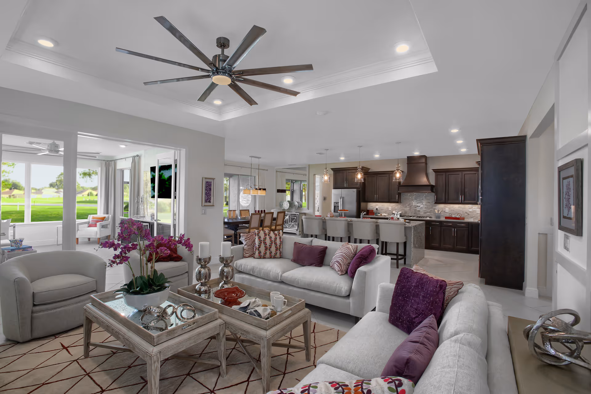 Open-plan living room and kitchen with gray sofas, wooden coffee tables, purple accent pillows and a large ceiling fan.