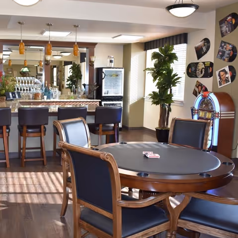 A well-lit common area with a round poker table and four chairs in the foreground. Behind it is a bar counter with four high chairs, pendant lights, and a mirrored wall. To the right, there is a jukebox and wall decorations featuring vinyl records and photos. Large windows with blinds and a potted plant add natural light to the space.