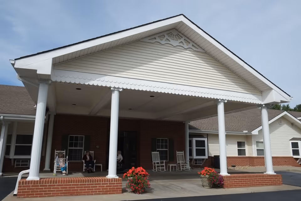 Front entrance of Hillside Village Of De Soto facility with a covered porch supported by white columns, brick base, and rocking chairs. There are a few elderly people sitting on the porch and flower pots with red flowers near the entrance.