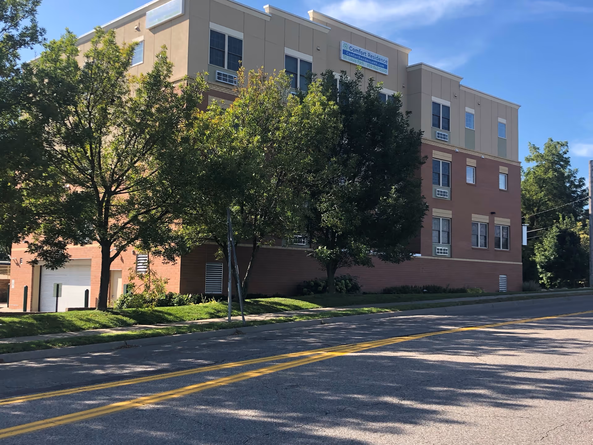 Multi-story brick and tan senior living building behind trees with a sidewalk and a street in the foreground.
