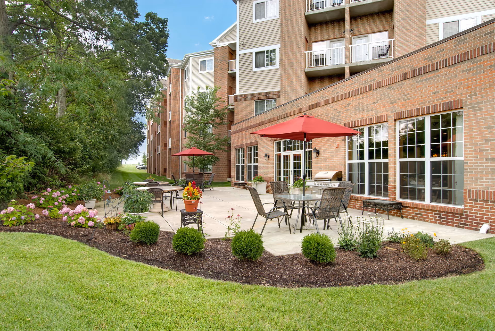 Outdoor patio area at Barrington of West Chester with tables and chairs under red umbrellas, surrounded by landscaped bushes, flowers, and green grass next to a brick building with large windows.