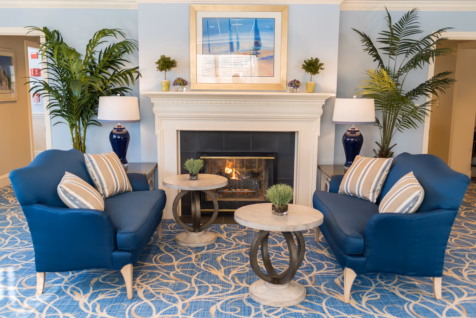 Cozy lounge area with two blue armchairs facing a fireplace, round side tables, lamps, and decorative plants.