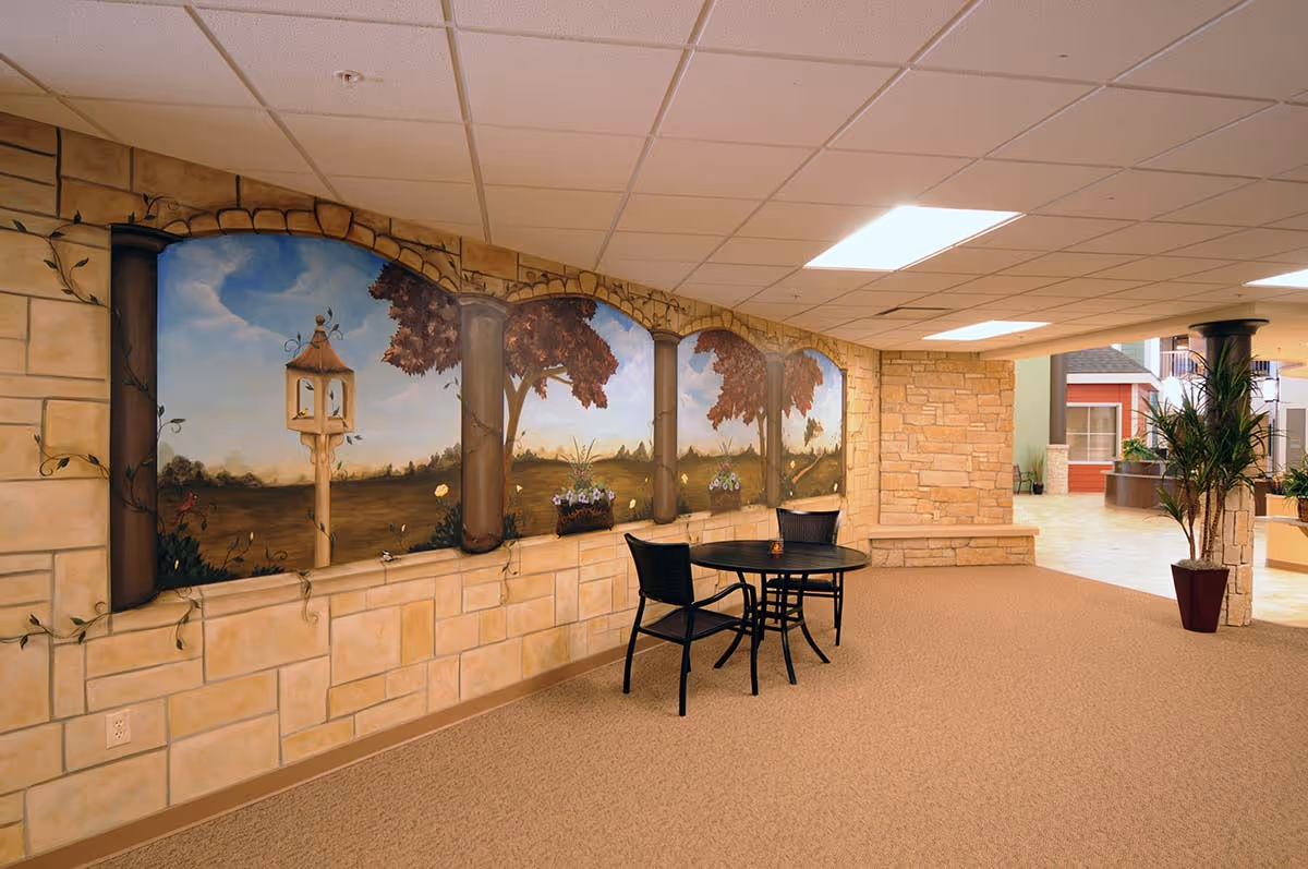 Indoor seating area with a round black table and two black chairs placed against a wall featuring a mural of a scenic landscape with trees, a birdhouse, and flowers. The room has beige carpet, stone-textured walls, and a ceiling with recessed lighting. There are potted plants and a view into another part of the facility with more seating and architectural elements.