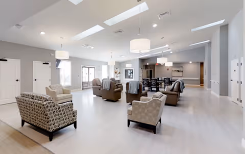 A spacious, well-lit common area with multiple armchairs and sofas arranged for socializing. The room features large windows, modern ceiling lights, and light-colored flooring. In the background, there is a dining table with chairs and a hallway leading to other rooms.