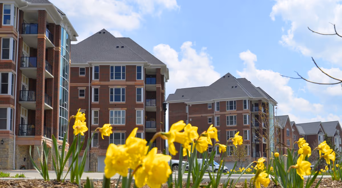 View of a senior living facility with multiple brick buildings under a partly cloudy sky. Yellow daffodil flowers are in the foreground, with a few cars parked near the buildings.