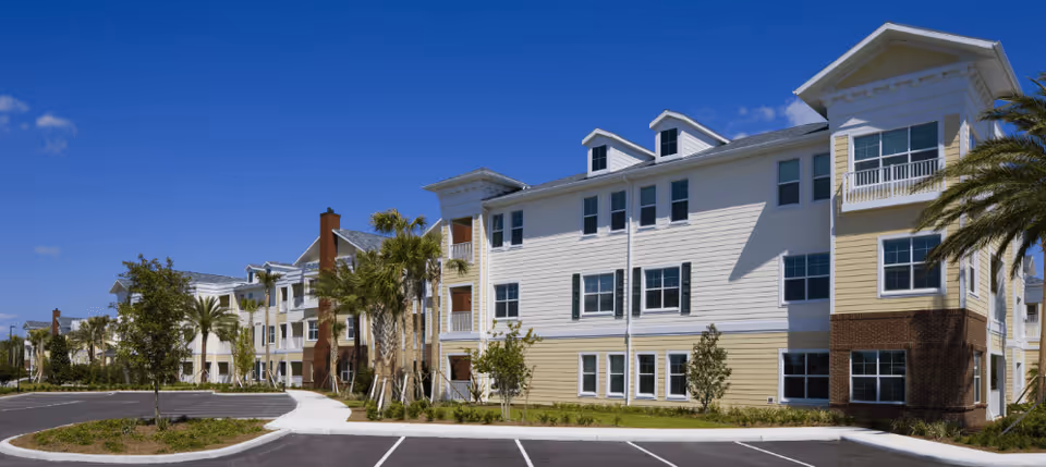 Exterior view of a multi-story senior living facility building with white and beige siding, several windows, palm trees, and a clear blue sky. There is a parking lot and landscaped area in front of the building.